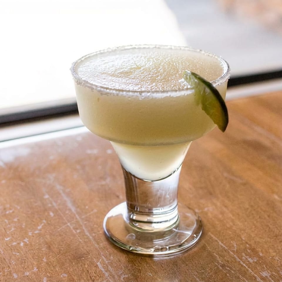 Pale green cocktail in a stemmed glass, garnished with a lime wedge, sits on a wooden table beside a window.