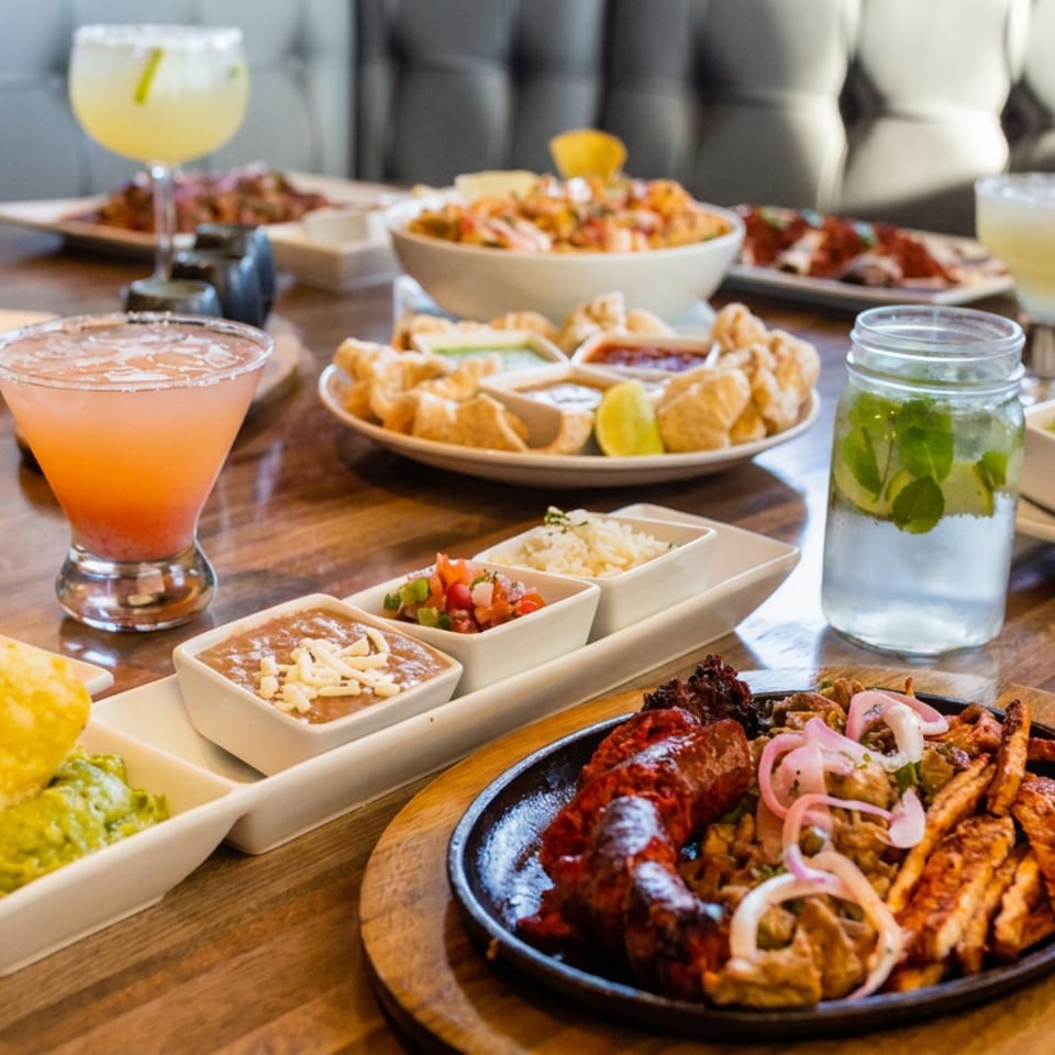 Restaurant table filled with Latin-inspired dishes, colorful appetizers, grilled meats, dipping sauces, and citrus cocktails.